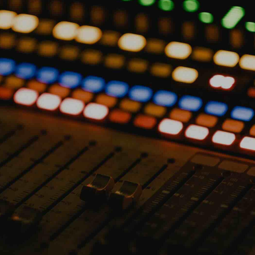 Close-up of an audio mixing console with faders in the foreground and colorful illuminated buttons blurred in the background.